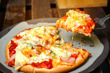 Freshly baked pizza hot from the oven. Slice of hot pizza lunch large cheese topped with tomato sauce and bell pepper vegetables. Italian fast food on a wooden table in side view.の写真素材