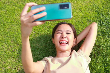 Beautiful smiling Asian woman, cheerful and happy. Lying on the green grass holding a smartphone to take a selfie. Concept of modern communication technology. social mediaの写真素材