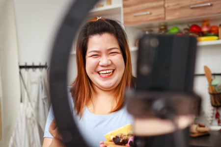 Fat Asian woman cooks organic food in her kitchen at home. Record online video via smartphone Make content published on social media. Influencer concept. healthy cooking for weight lossの写真素材