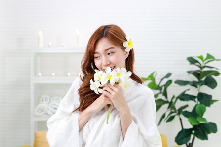 Portrait of a beautiful Asian woman doing a spa at home smiling happily. Dressed in white, standing holding flowers. Asian beauty conceptの写真素材