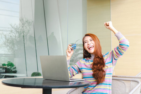 Asian woman sitting at work and holding credit card Arms raised, happy, smiling, happy. online shopping conceptの写真素材