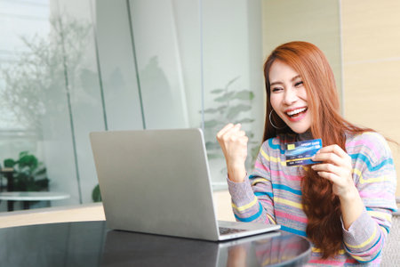 Asian woman sitting at work and holding credit card Arms raised, happy, smiling, happy. online shopping conceptの写真素材