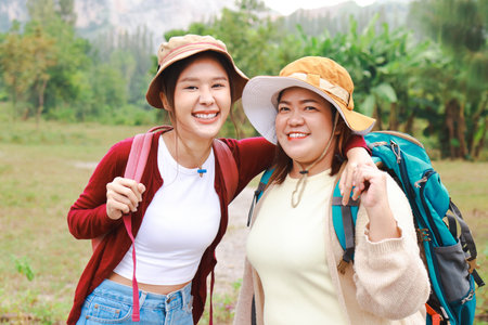 Portrait of two Asian female travelers carrying their backpacks, hugging their necks, smiling cheerfully, enjoying nature travel. Travel concept. Tourists hiking, camping.の写真素材