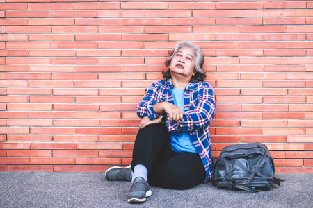 Homeless elderly woman sitting on the floor of the corridor back against brick wall Stressed out with life. copy spaceの写真素材