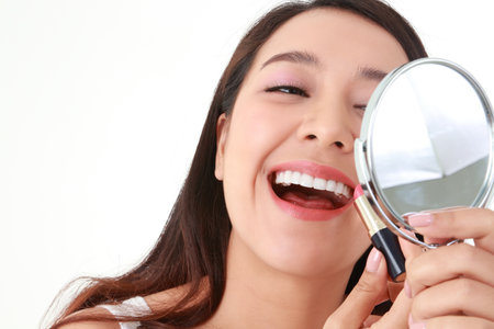 Beautiful smiling Asian woman applying lipstick to her mouth, holding a mirror to her face. Beauty concept. Asian woman with beautiful skin. Soft and smooth. Healthy. White background.の写真素材