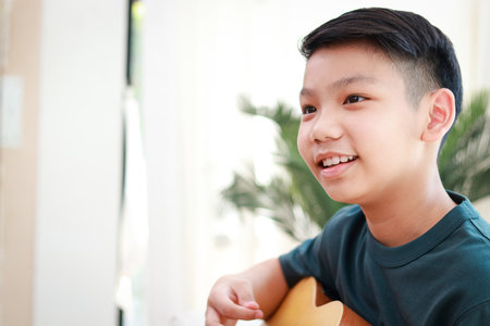 Portrait of Asian boy playing acoustic guitar music sitting on sofa in house. Music concept. Child learning.の写真素材