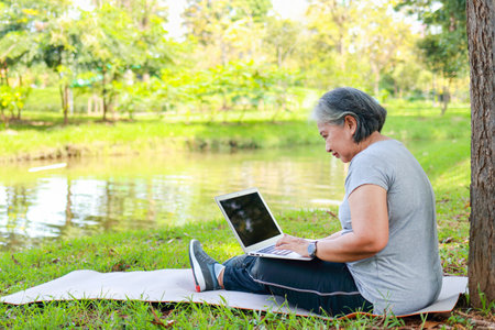 Asian senior woman sitting at the park working with laptop computer. life in retirement. Copy spaceの写真素材