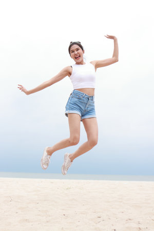 Cheerful young beautiful Asian woman having fun in white shirt and shorts jumping high on the beach. Smiling, happy to travel to the sea on weekends. travel conceptの写真素材