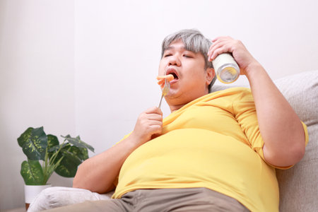 Fat Asian man eating hot dogs and beer on the sofa in his room at home. Health concept, weight lossの写真素材