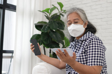 Asian elderly woman in face mask sits at home sofa holding smartphone talking with doctor online.の写真素材