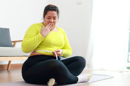 Fat asian woman exercising at home using a meter tape to measure her waist circumference. She looks shocked at the size. Sports concept, health care. weight lossの写真素材
