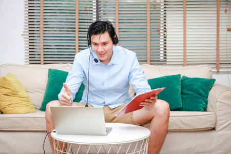 Asian male businessman working from home using laptop computer and wearing headphones with microphone meeting online work.の写真素材