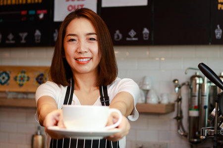 Asian female worker holding a mug of hot coffee in a coffee shop, she smiles happily enjoying her work. Entrepreneurial concept. restaurant businessの写真素材
