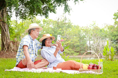 Happy Asian elderly couple Picnic sitting in the park watching social media on smartphones together. Living in retirement to be happy. Elderly health careの写真素材