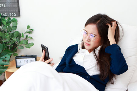 Asian woman in suit lying on bed holding smartphone online chatting work She has stress. Female employees work overtime. Business concept, Entrepreneurの写真素材