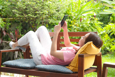 Asian woman resting on a bench at home watching social media on her smartphone. Living life to be happy. health careの写真素材