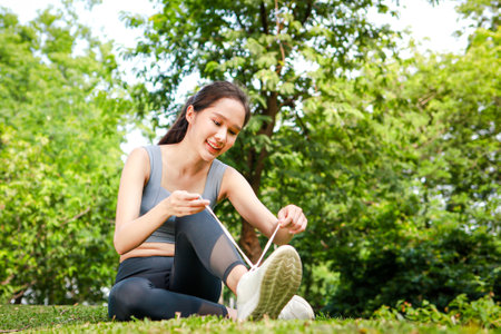 Beautiful Asian woman exercising in the park, she sits on the grass tying her shoelaces. Sport...