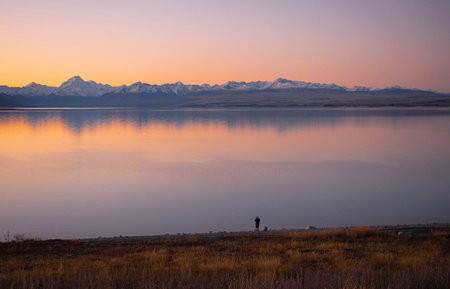 Person on lakeshore taking a photo of mountain mirror reflection panorama at sunset, Southern Alps, Mackenzie, Canterbury, South Island, New Zealandの写真素材