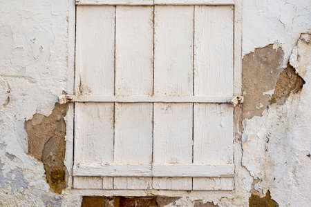 White cracked wall with a peeling paint with a window closed with a painted white wooden shutters and locked with two locksの写真素材