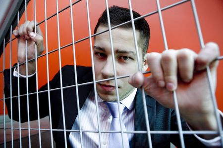 Portrait of young agressive man in business clothes behind bars with red backgroungの写真素材