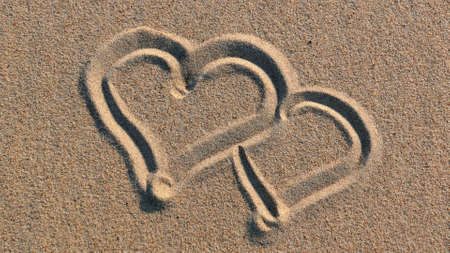 Drawing Heart Shapes on Sandy Beach with Wooden Stick at Sunsetの写真素材