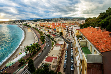 Amazing sunset view on red tile roofs of Nice town and dramatic sky. Cars on auto road which along coastline (Promenade de Anglais and wide pedestrian zone for jogging, moving on a board, on rollers. View from Colline de Chateau (hill)のeditorial素材