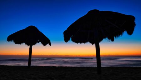 Golden and blue sunrise with straw umbrellas in backlight on a deserted tropical beach with the Caribbean sea in the backgroundの写真素材