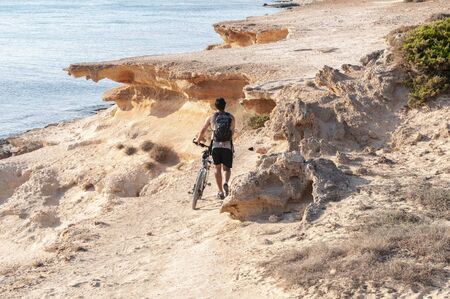 An unknown tourist walks with his backpack along a path overlooking the sea with his bicycle. Formentera island, Mediterranean sea, Spainの写真素材
