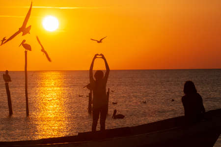 Girls watch the sunset on a fishing boat near the beach. Seabirds flying. Girl with raised arms, hands in heart symbol. In the background the orange sky and the sun on the horizon. Holbox Islandの写真素材