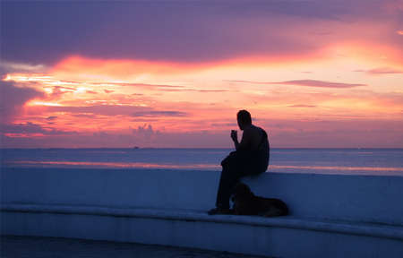 Silhouette of a man watching the sunset accompanied with his dog. Unknown man observes the sunset in Cozumel, Mexico. In the background the Caribbean sea and the sky with colored cloudsの写真素材