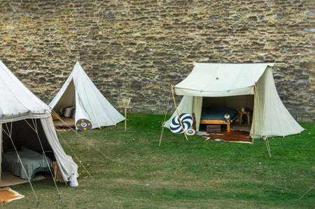Medieval scene against the historical backdrop of an old city wall, village with tents of various designs, view of tents, village square with utensils and containers, furniture, wooden storage and transport cartのeditorial素材
