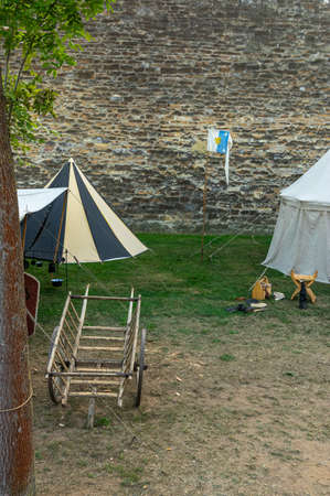 Medieval scene against the historical backdrop of an old city wall, village with tents of various designs, view of tents, village square with utensils and containers, furniture, wooden storage and transport cartのeditorial素材