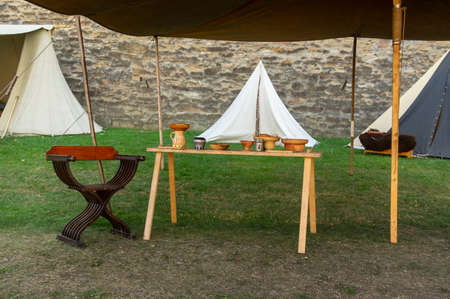 Medieval scene against the historical backdrop of an old city wall, village with tents of various designs, view of tents, village square with utensils and containers, furniture, wooden storage and transport cartのeditorial素材