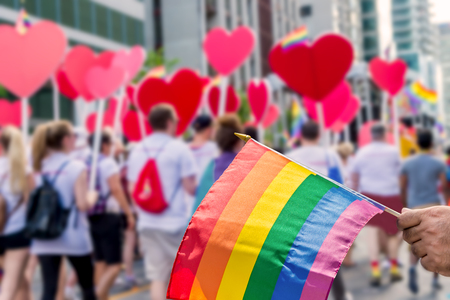 Rainbow flag floating in front of blurred gay pride participants holding heartsの写真素材