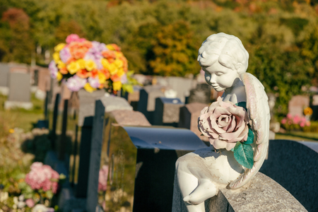 Statue of an angel holding a rose sitting on a tombstone in a cemetery with flowers and headstones in the backgroundの写真素材