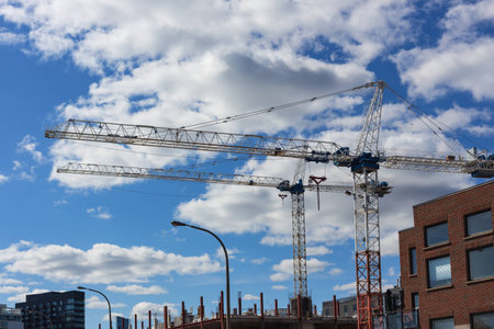 two tall cranes on construction site against the blue sky and clouds with buildings and street lamps