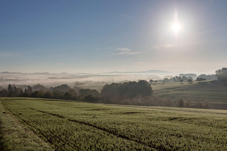 rural landscape agriculture field in foreground heavy fog mist on fields and village in middle and hills and wind turbines in background against cloudy blue skyの写真素材