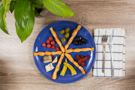 top view mediterranean antipasti plate with two kinds of green black olives fresh dried tomatoes cheese filled peppers separated by breadsticks on blue plate napkin cutlery pot plant on wooden tableの写真素材