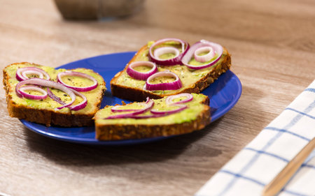 three avocado toast seasoned with salt and pepper and red onion cut in rings on a blue plate with napkin and cutlery on a wooden tableの写真素材