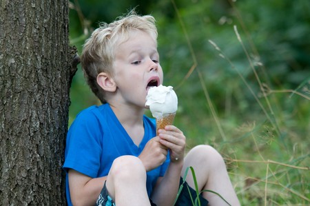 Seven year old boy enjoys eating a big icecream on a hot summerday, while sitting against a tree.の写真素材