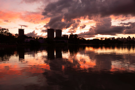 Dramatic orange sky reflected in the calm waters of the lake, while the silhouettes of buildings and trees create an urban setting at dusk.の写真素材