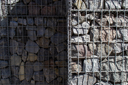 Gabion wall structure featuring wire mesh cages filled with irregularly-shaped stones. The metal...