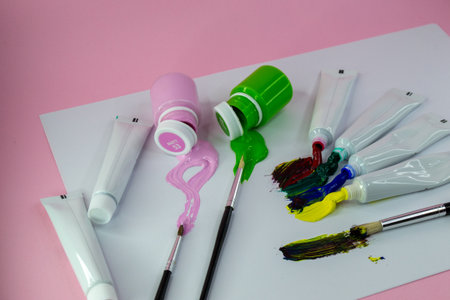 Various bright oil paints, including pink, green, yellow, and blue, are spread on white paper. Plastic bottles and tubes sit casually, some open, with paint brushes nearbyの写真素材