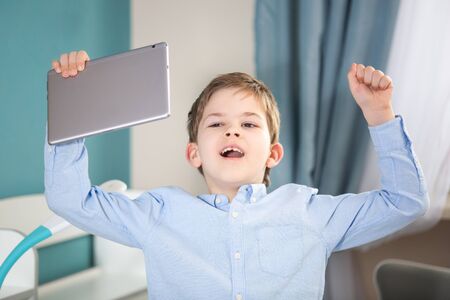 technology, learn form home - lovely small boy in blue shirt jumping near his desk holding his tablet and rejoycingの写真素材
