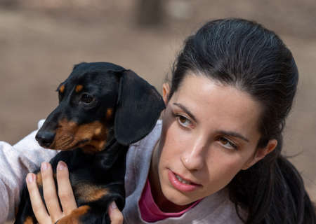 Young caucasian woman hugging black smooth haired miniature dachshund in forest