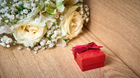 Red gift box with bow on lid, bouquet of white flowers on wooden background, holiday.の写真素材