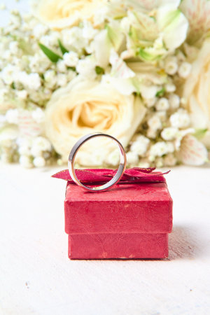 A silver ring stands on a red gift box and a bouquet of flowers on a white textured tree background, holiday.の写真素材
