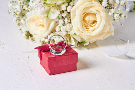 Two wedding rings of different sizes, a red gift box and a bride's bouquet lie on a white wooden background.の写真素材