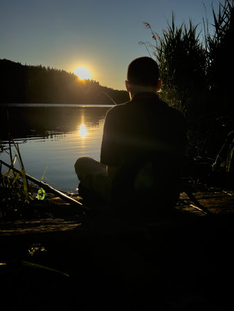 Young man fishing on a quiet lake during sunset. A quiet moment of solitude and relaxation in nature at the water's edgeの写真素材