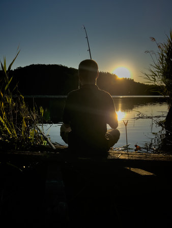 Young man fishing on a quiet lake during sunset. A quiet moment of solitude and relaxation in nature at the water's edgeの写真素材